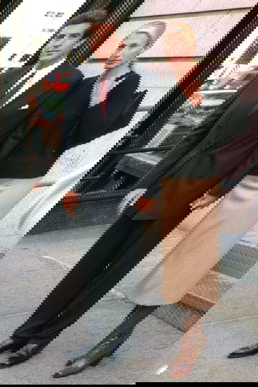 Пара держится за руки на улице у городского здания: деловой стиль мужчины и лаконичный наряд женщины, походный снимок папарацци.