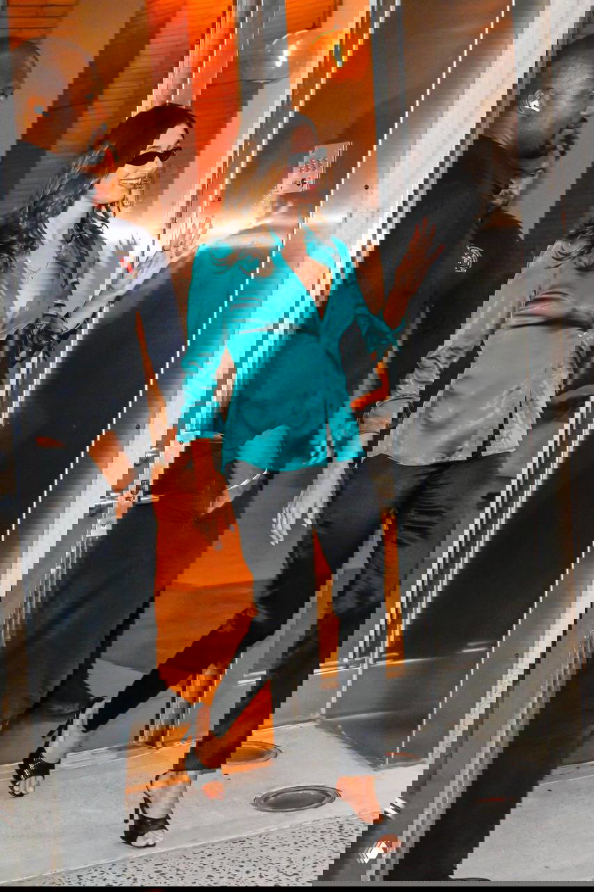 Женщина в атласной зелёной рубашке и чёрных брюках выходит из здания вечером, образ в стиле стритстайл с туфлями на каблуке.