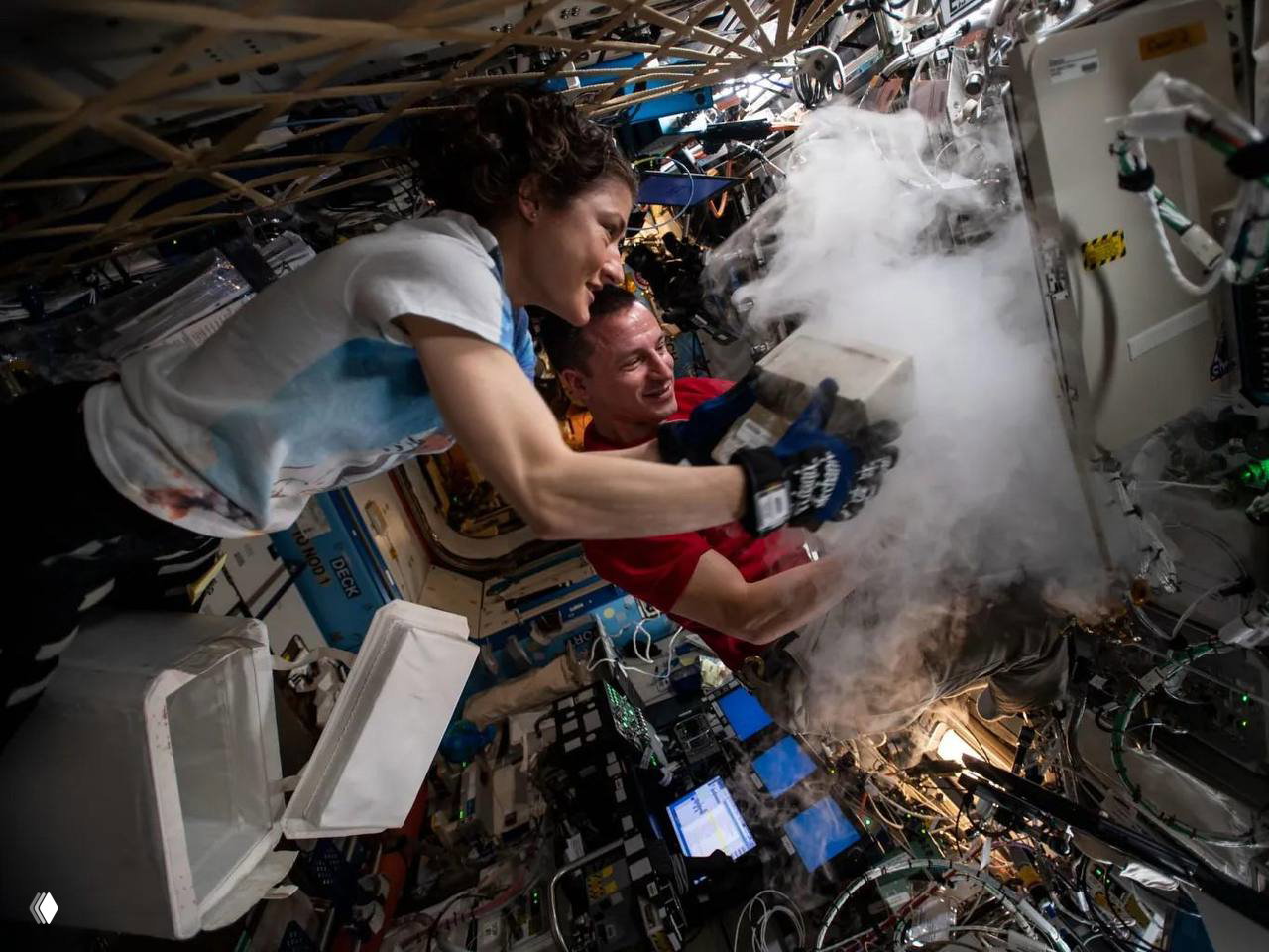 Кристина Кук внутри модуля МКС выполняет эксперимент в условиях невесомости, видны приборы, оборудование и другой член экипажа.