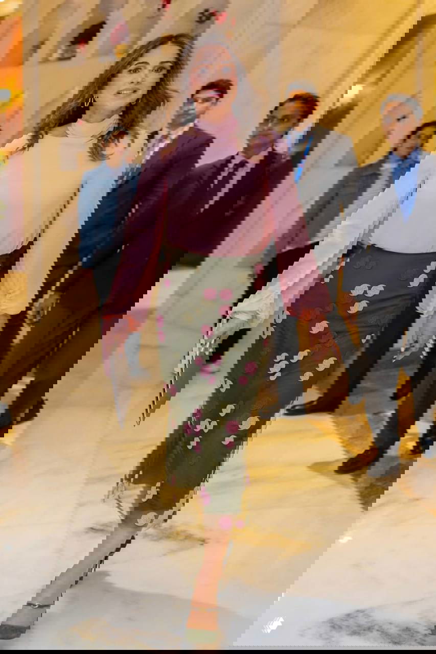 Женщина в нежно-розовой блузке и оливковой юбке с декоративными кружками идёт по коридору отеля в окружении сотрудников протокола и охраны.