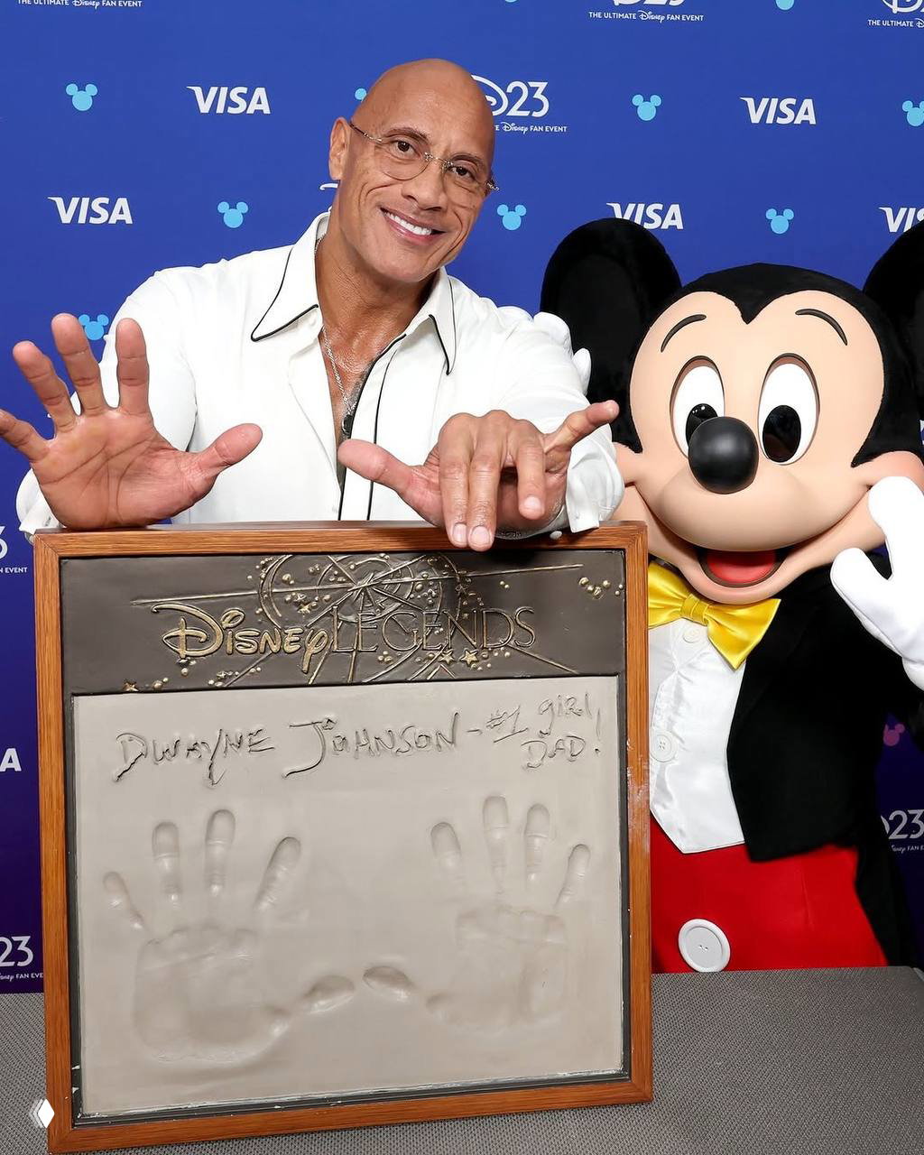 Дуэйн Джонсон улыбается у плакетки Disney Legends с отпечатками рук и надписью, рядом виден Микки Маус на фоне пресс‑зоны и логотипов D23