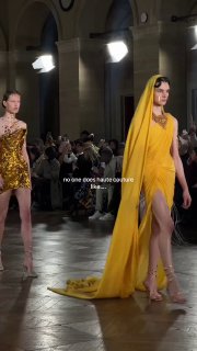 Модель в длинном жёлтом платье с шлейфом идёт по подиуму на показе Gaurav Gupta; вокруг зрители и фотографы в зале под светом софитов.