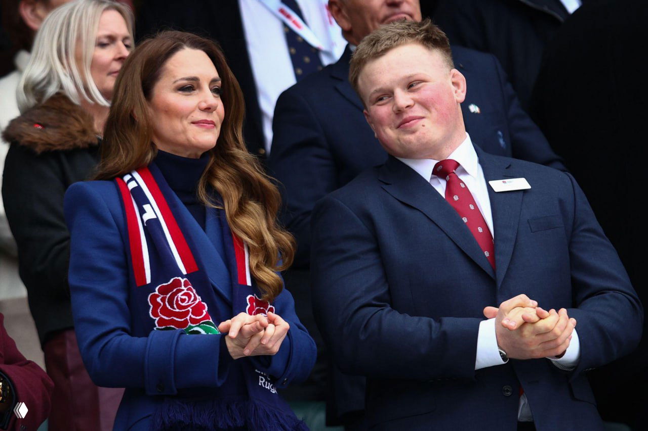 Женщина в синем пальто и фанатском шарфе England Rugby аплодирует рядом с мужчиной в костюме на стадионе; кадр с трибуны.