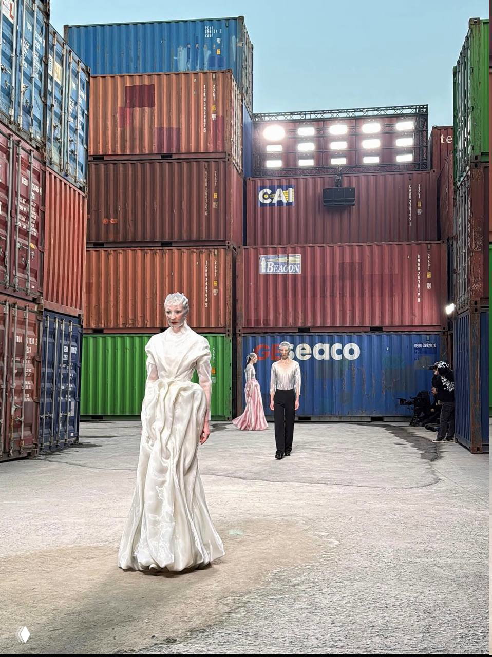Модели шагают по проходу между высокими контейнерами: белые и пастельные наряды на фоне индустриальной сцены шоу Maison Margiela в Шанхае.