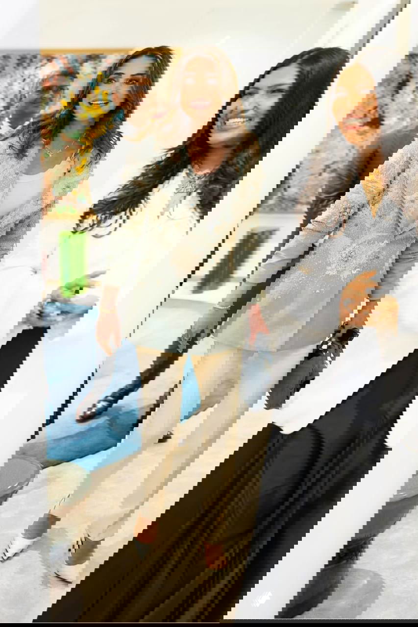 Женщина в вышитом светлом жакете и бежевых брюках улыбается во время приёма в галерее; вокруг гости и элементы современного интерьера.