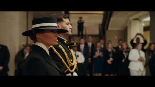 Кадр из трейлера фильма: Мелания Трамп в чёрном наряде и шляпе наблюдает церемонию инаугурации, люди и охрана на заднем плане.
