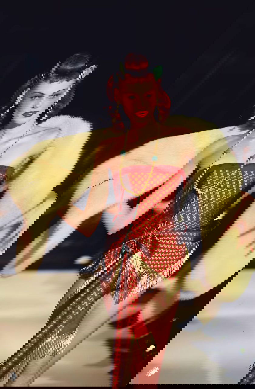 Модель в корсетном красном наряде и светлой меховой накидке на подиуме Dior осень‑зима 1997, бахрома, драпировка и ретро‑атмосфера шоу.