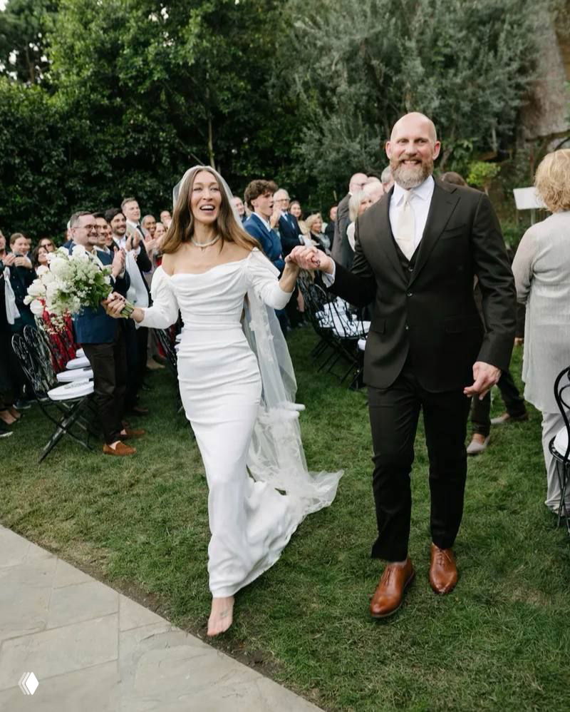 Невеста в белом платье идёт по проходу рука об руку с женихом в тёмном костюме — кадр со свадебной церемонии на открытом воздухе среди гостей.