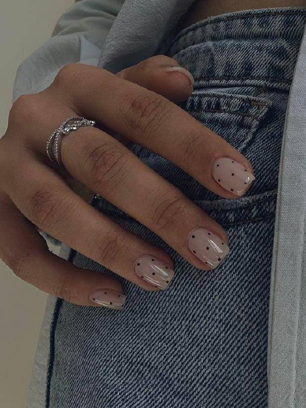 Крупный план руки на джинсах с короткими прозрачными ногтями и мелкими тёмными точками — мягкий миндаль/овальная форма с декоративным дизайном.