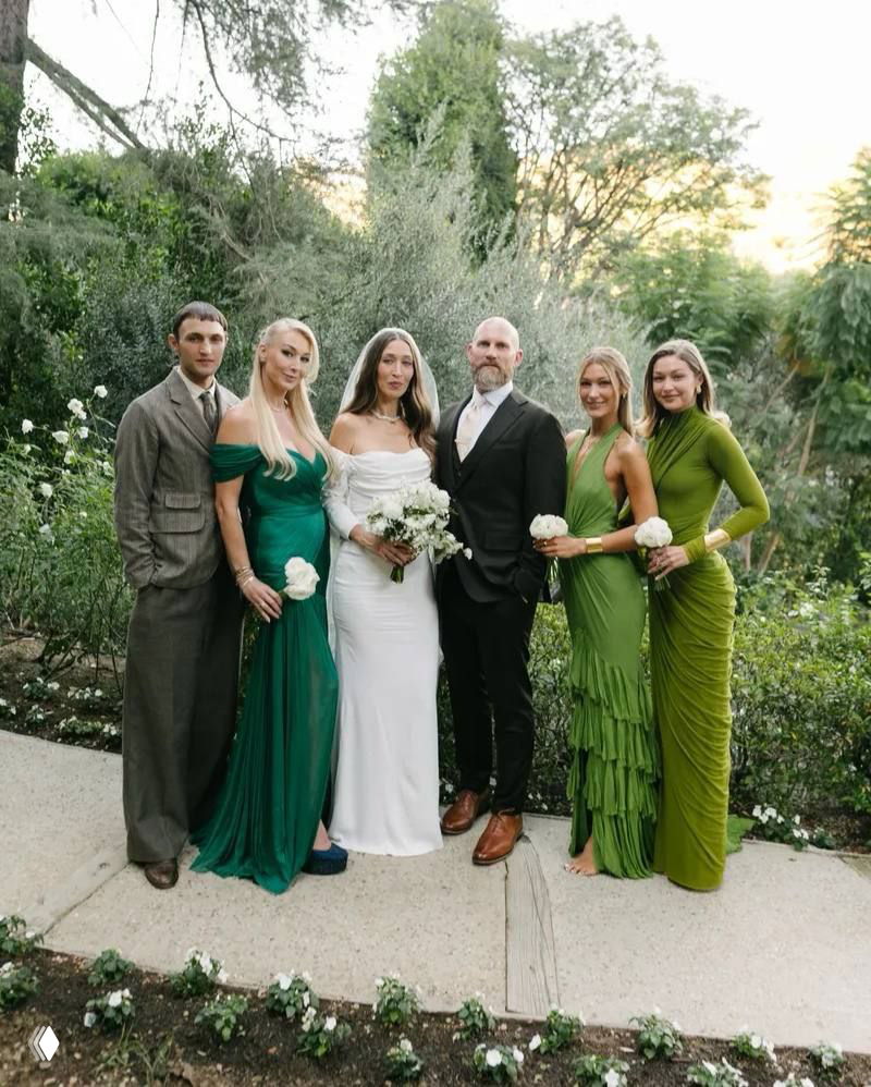 Групповой портрет на природе: невеста в белом, жених и рядом гости в разных оттенках зелёного на фоне садового ландшафта и цветочных клумб.