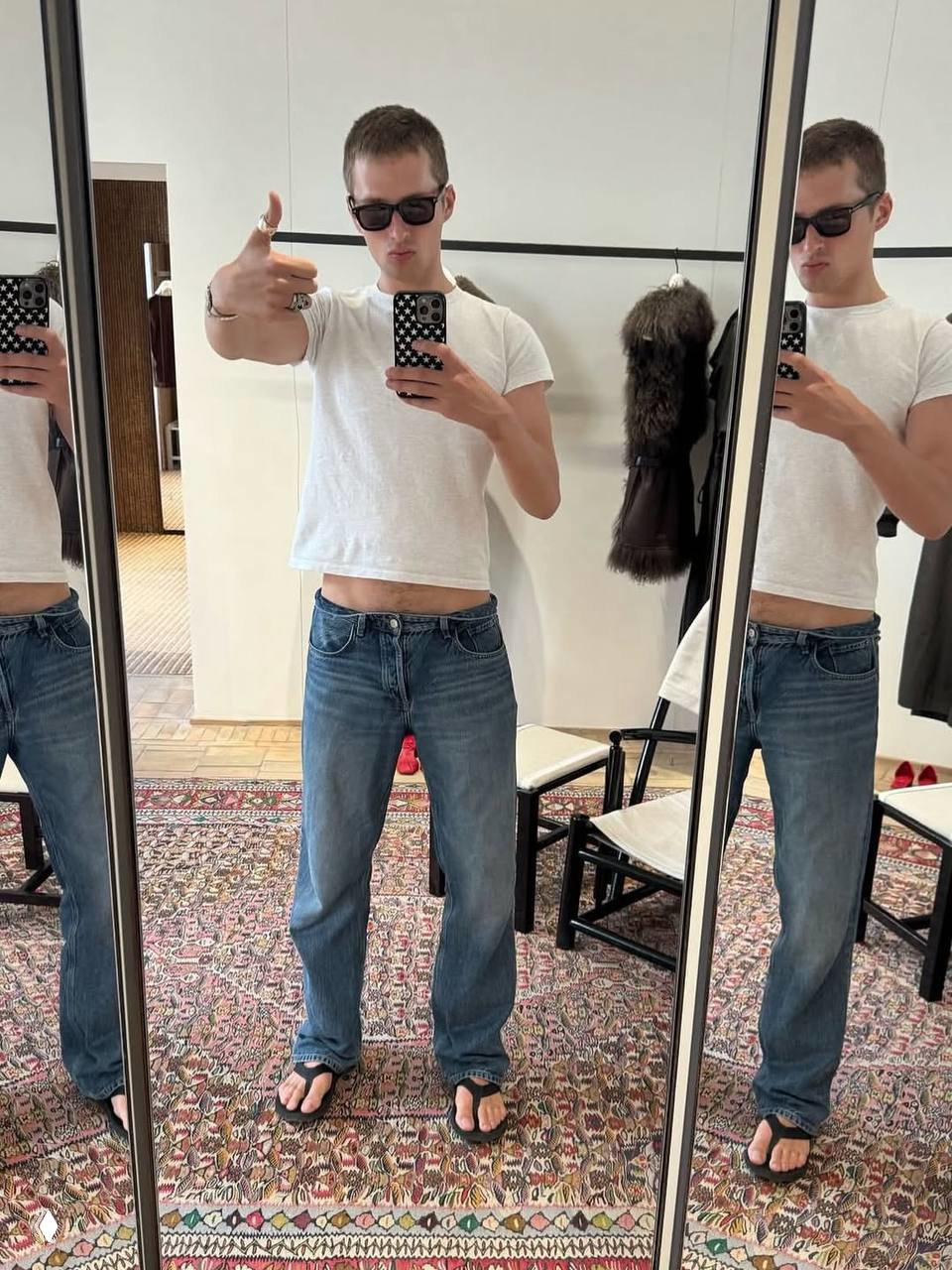 Зеркальное селфи мужчины в белой футболке и джинсах в примерочной или бутике, отражения в трех панелях зеркала, повседневный стиль.