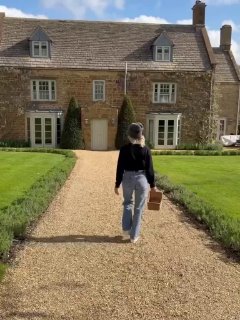 Памела Андерсон идёт по гравийной дорожке к старинному дому-усадьбе, видна спина, джинсы и плетёная корзинка в руках.