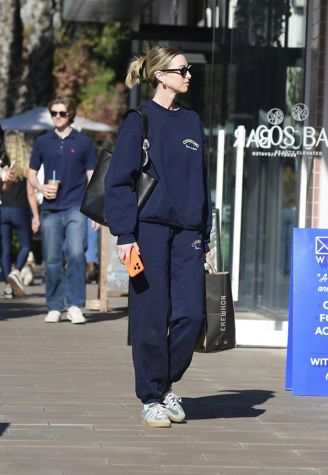 Фигура женщины в синем свитшоте и штанах на тротуаре; в руках смартфон и шоп‑пакет, образ выглядит уютно и расслабленно.