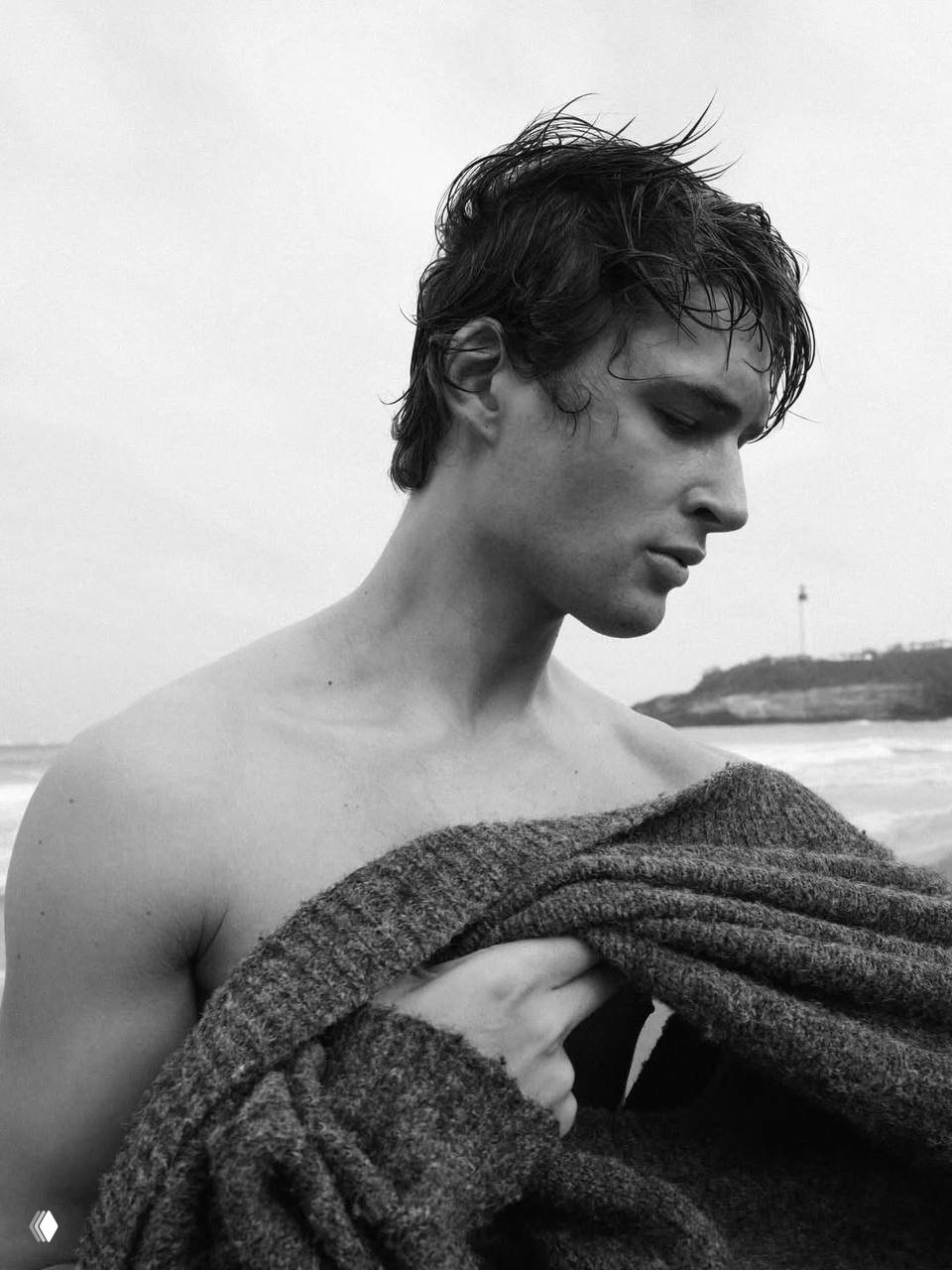 Портрет в профиль у моря: мужчина накрывает тело джемпером, черно‑белая эстетика модной съёмки, крупный план лица и плеча.