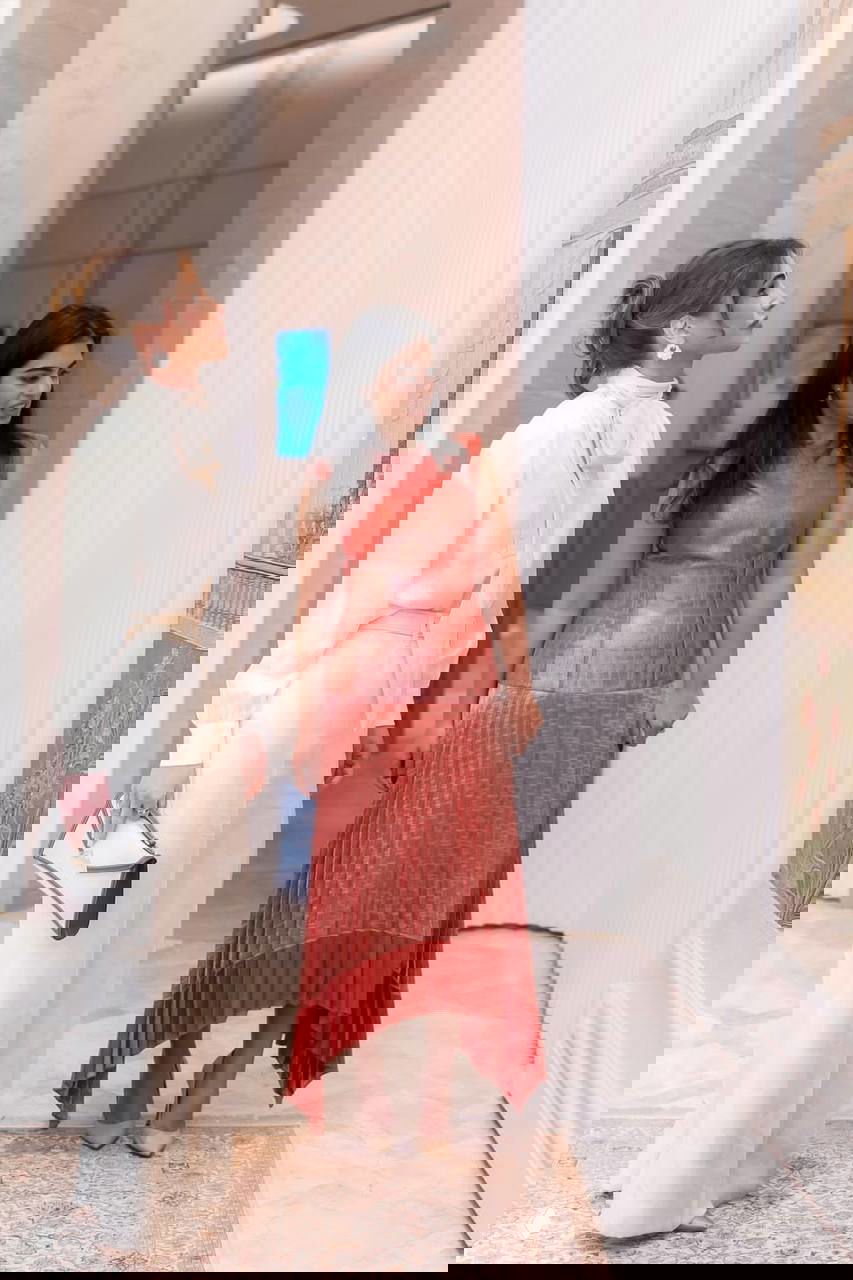 Две женщины внимательно рассматривают экспонаты в светлом выставочном зале; одна в молочном брючном костюме, другая в красном платье, интерьер с ковровым покрытием.