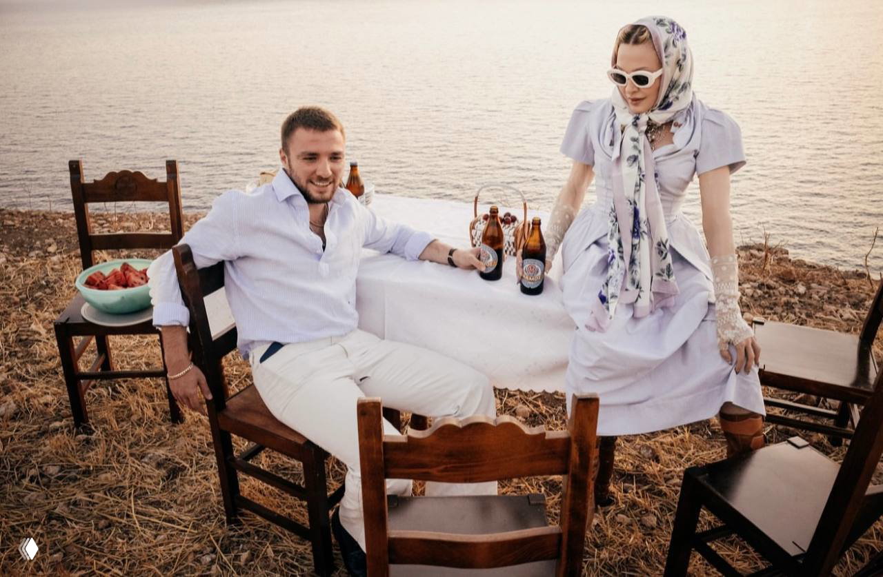 Два человека сидят за столом на фоне моря и скалистого берега; вечерний свет, бутылки и тарелки на столе создают атмосферу отдыха.