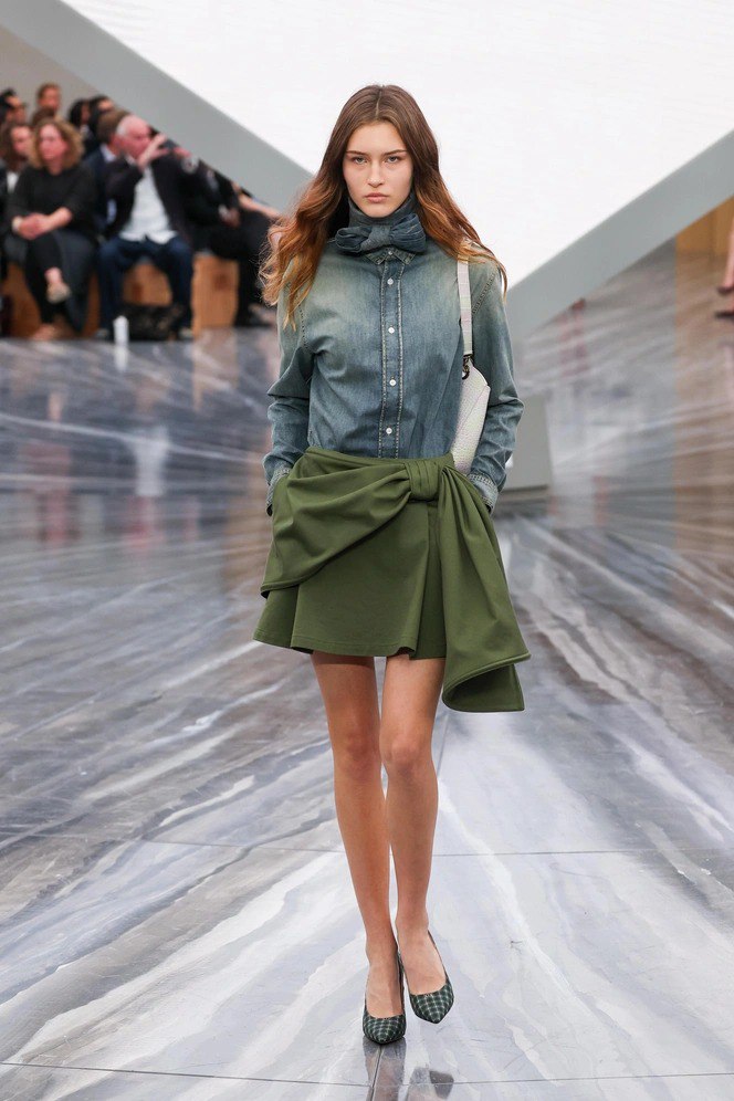 Модель в джинсовой рубашке и мини‑юбке в нейтральных тонах на глянцевом подиуме, подчёркнуты пастельные акценты и минимализм кроя.