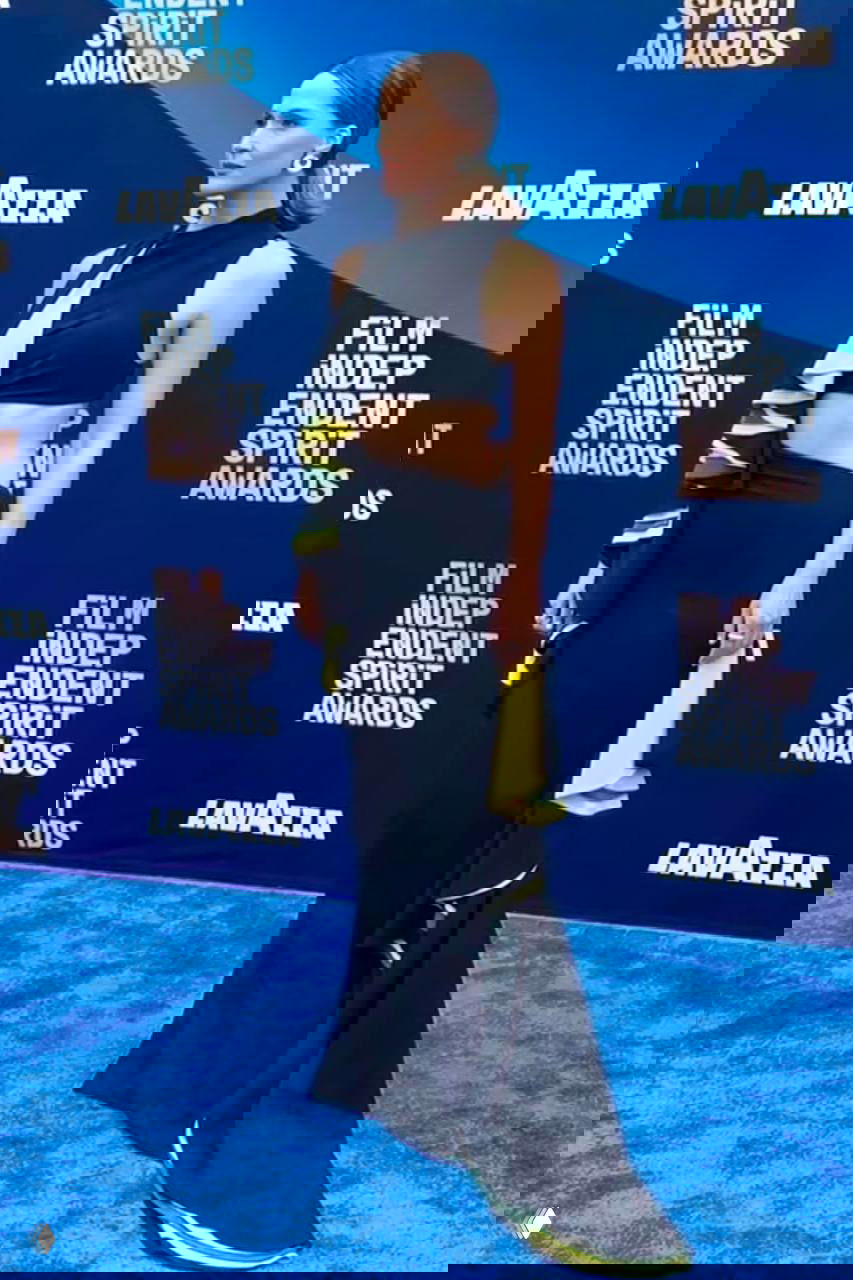 Гостья в чёрном платье с вырезами и яркими цветными акцентами на синем фоне фотозоны; профильный ракурс, видно украшения.