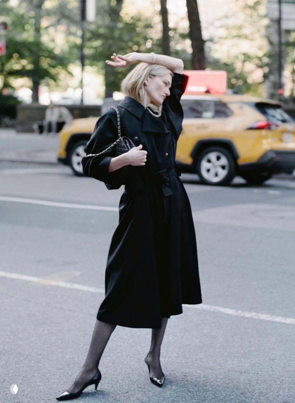 Сара Пиджон в длинном чёрном плаще идёт по улице Нью‑Йорка на фоне жёлтого такси, уличный образ в движении и городской антураж.