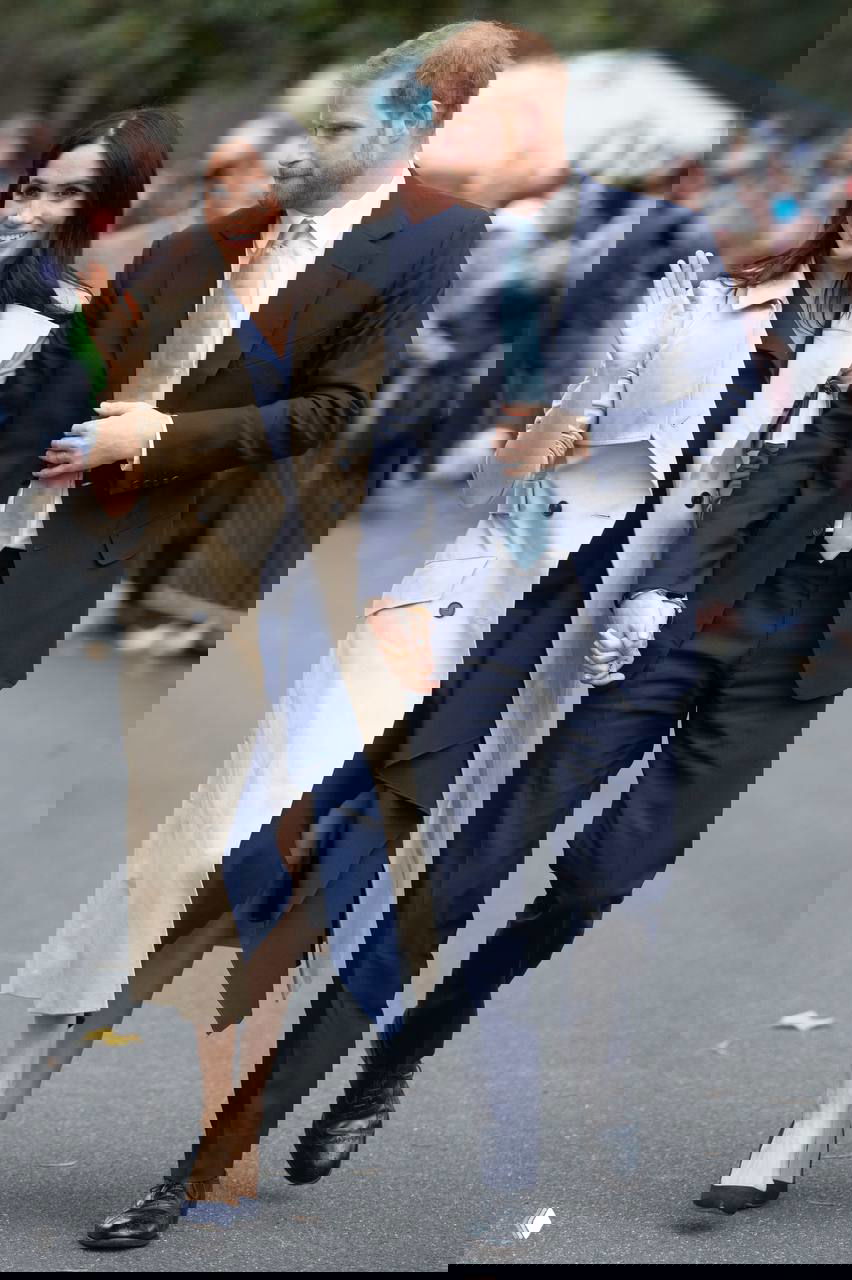 Уличный снимок: пара идёт по городу, женщина в бежевом тренче и мужчина в костюме; городской фон и прохожие подчёркивают повседневную элегантность плаща.