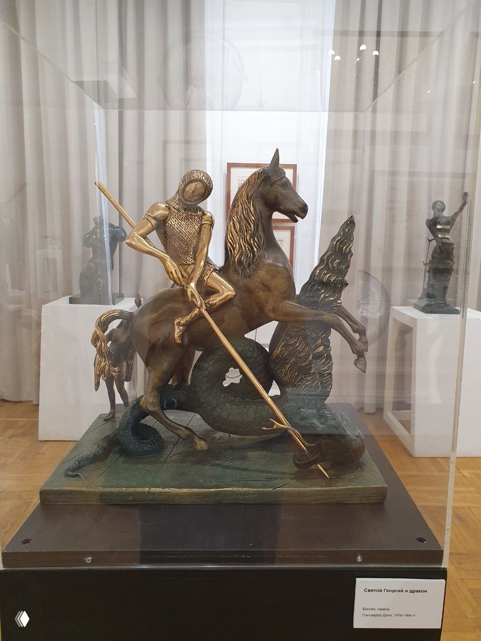 Бронзовая композиция всадника на лошади в витрине — символическая сцена с копьём и мифологическими мотивами, музейный фон просматривается сквозь стекло.