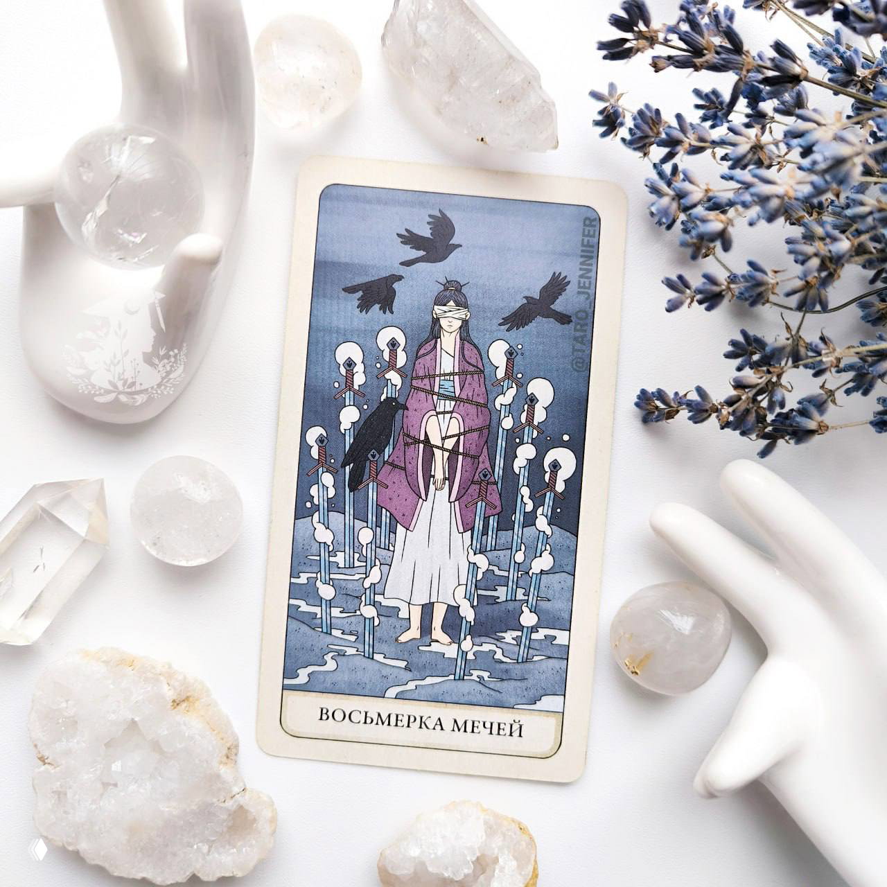 Крупный план карты «Восьмерка Мечей» из колоды Таро Четырех Дорог на белом фоне в окружении кристаллов, стеклянных шаров и ветки лаванды.