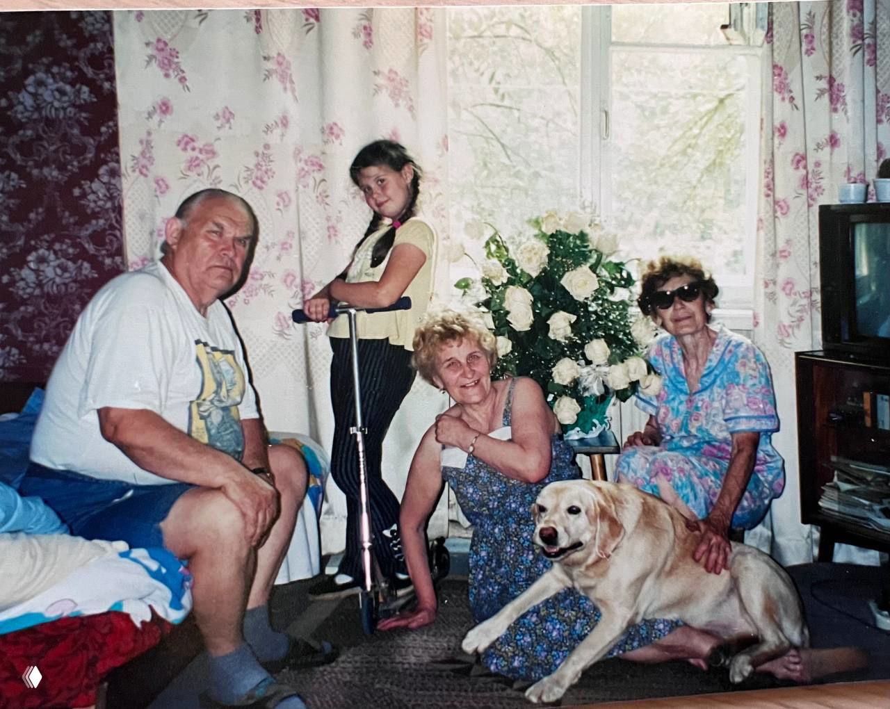 Семейный кадр: дети с бабушкой, дедушкой и собакой-лабрадором на фоне букета белых роз и занавесок — тёплый домашний момент.