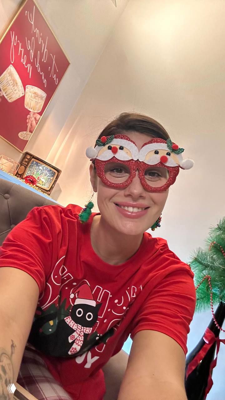 Селфи той же женщины в праздничных очках и красной футболке, видно елку и часть интерьера; радостное выражение лица и домашняя атмосфера.