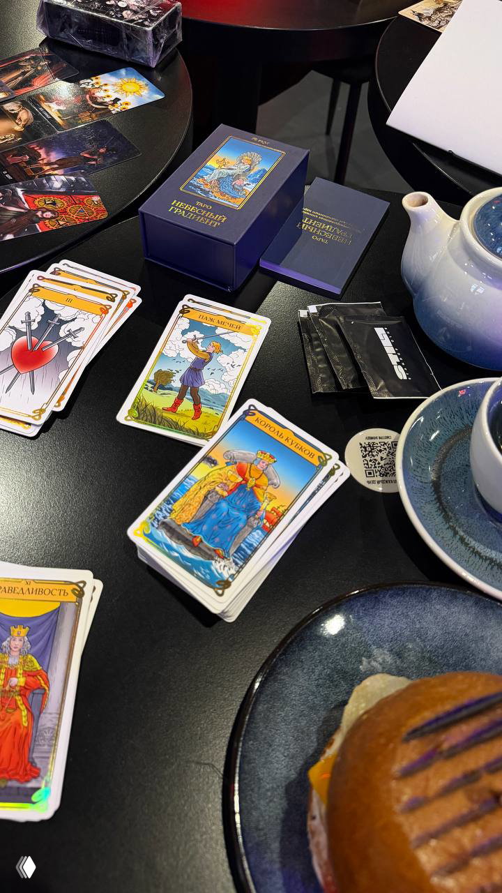 Фотография стола с раскладом Таро: несколько раскрытых карт, коробка колоды, чашка чая и булочка — атмосфера мастер-класса в женском клубе.