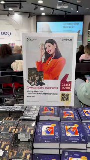 Фото крупного рекламного постера презентации с датой и QR‑кодом на фоне прилавка, где лежат коробки оракула и книги