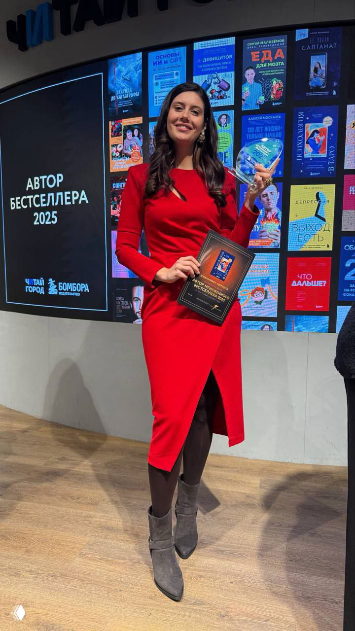 Дженнифер Митчелл в красном платье с наградой и дипломом на сцене книжного мероприятия, на заднем плане обложки книг