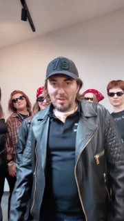 Кадр видео-приглашения: Роман Калинин в кожаной куртке перед камерой, за ним команда организаторов записывает обращение к участникам.