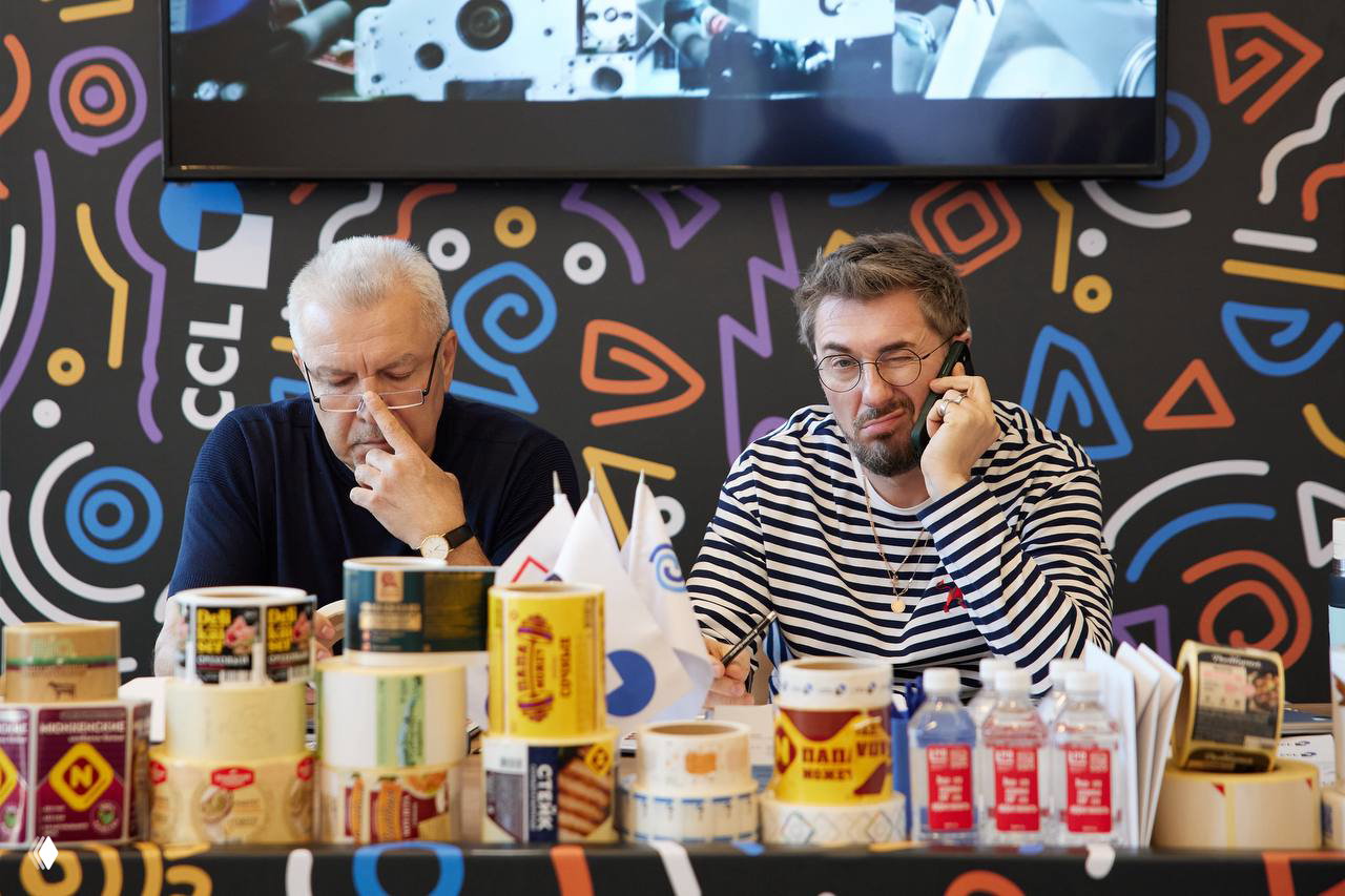 Выставочный стол с образцами упаковки и стикеров, два человека за стойкой занимаются организационными вопросами экспозиции на событии.