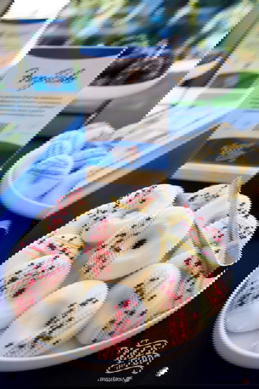 Блюдо с белой глазированной пончиковой продукцией на столе выставки, крупный план сладких изделий и щипцы для дегустации на фоне оформления.