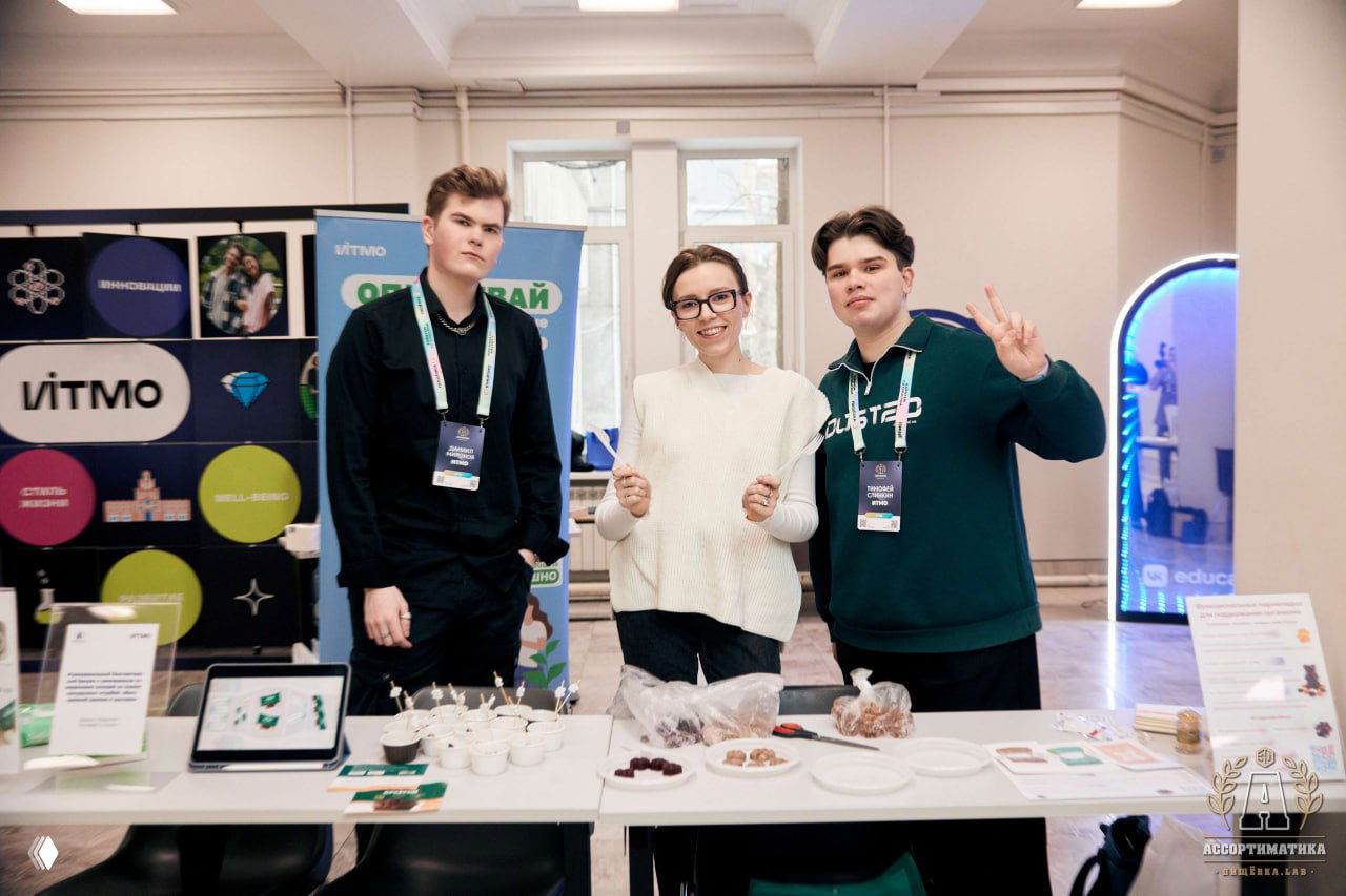 Трое молодых участников за выставочным столом на Ассортиматике в ИТМО: образцы продуктов, планшет, информационные материалы и улыбки.
