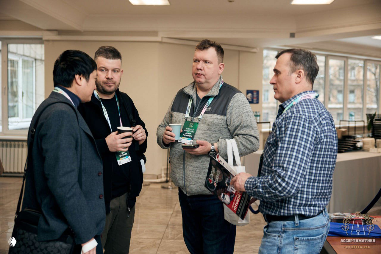 Группа участников конференции общается в холле с чашками кофе: обмен визитками, бейджи на шнурках и деловая неформальная беседа.
