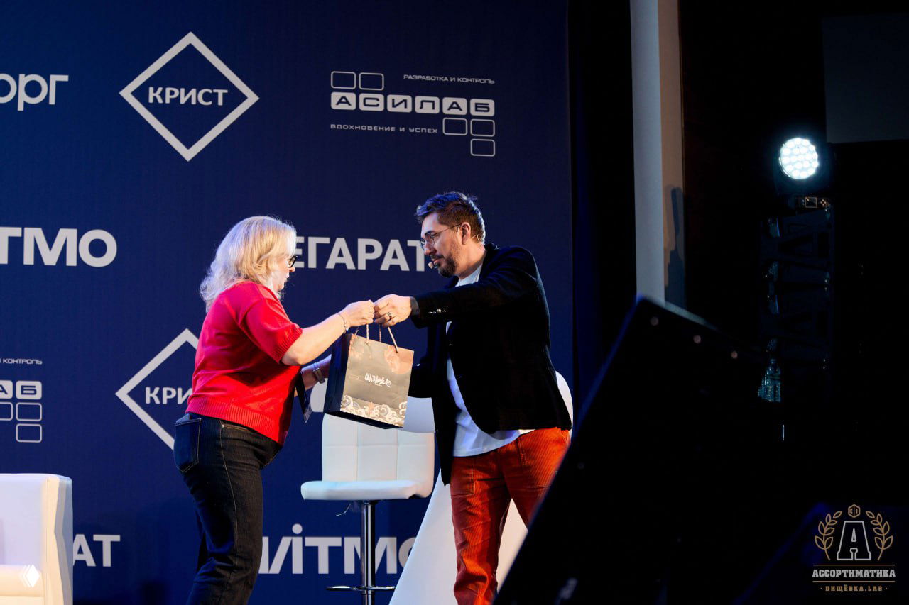 Вручение подарка на сцене: ведущий передает пакет участнице на фоне синего баннера с логотипами партнёров Ассортиматики.