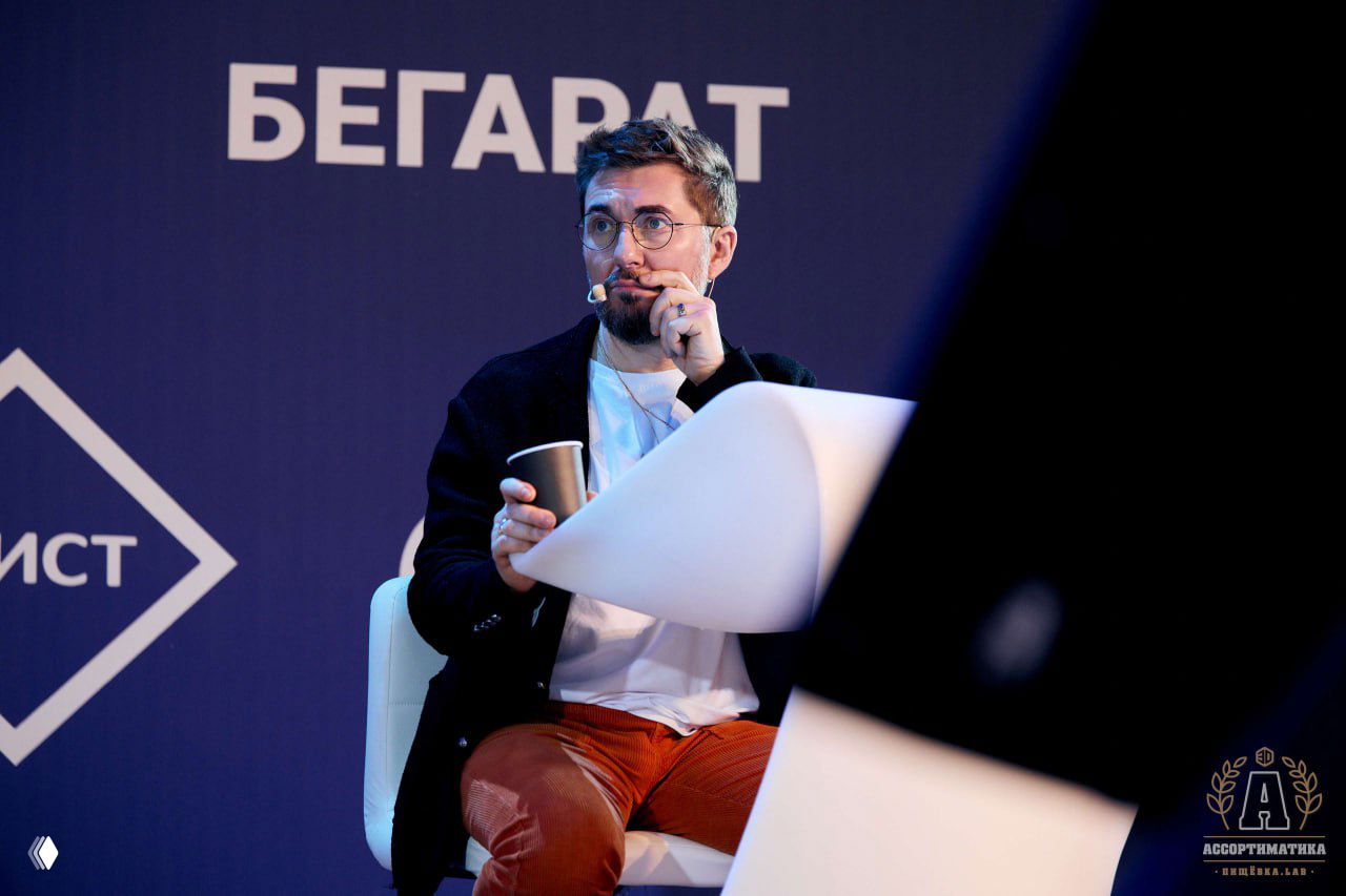 Близкий портрет спикера в кресле на сцене: микрофон-гарнитура, стакан кофе в руках и спокойная сосредоточенная поза во время паузы в дискуссии.