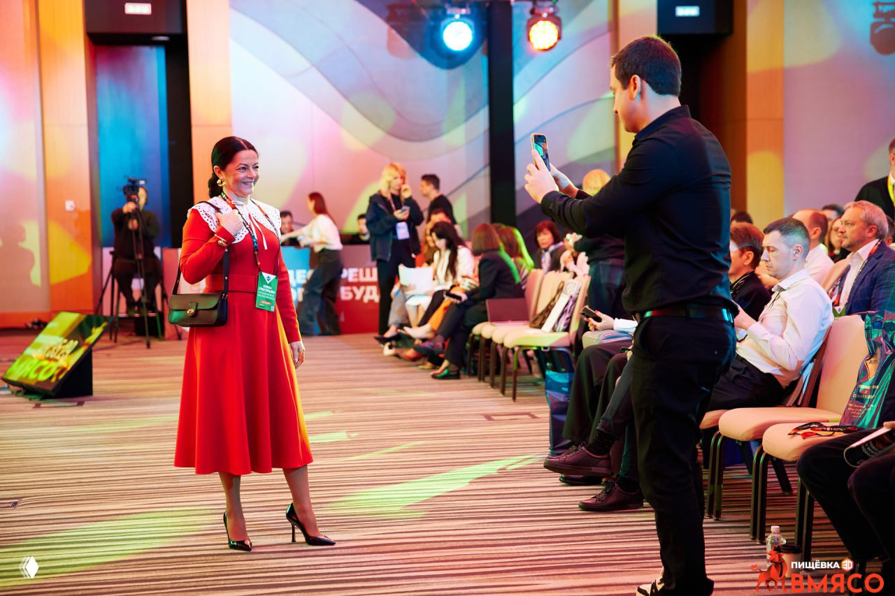 Эмоциональные кадры с мероприятия: женщина в красном платье проходит по ряду кресел, участники фотографируют и улыбаются под цветной подсветкой.