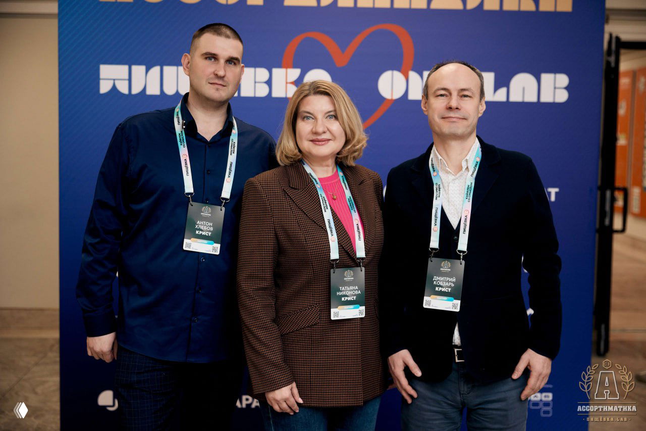 Трое организаторов или гостей позируют у бренд-стены Ассортиматики в ИТМО: бейджи, официальная фотозона и дружественная атмосфера.