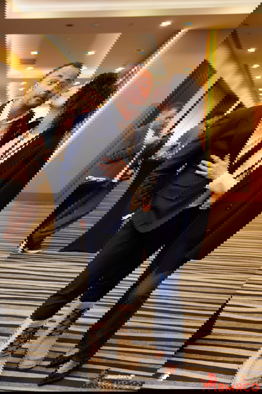 Неофициальная часть мероприятия: участники в коридоре обмениваются приветствиями, мужчина в костюме и женщина в деловом стиле демонстрируют тёплую атмосферу конференции.