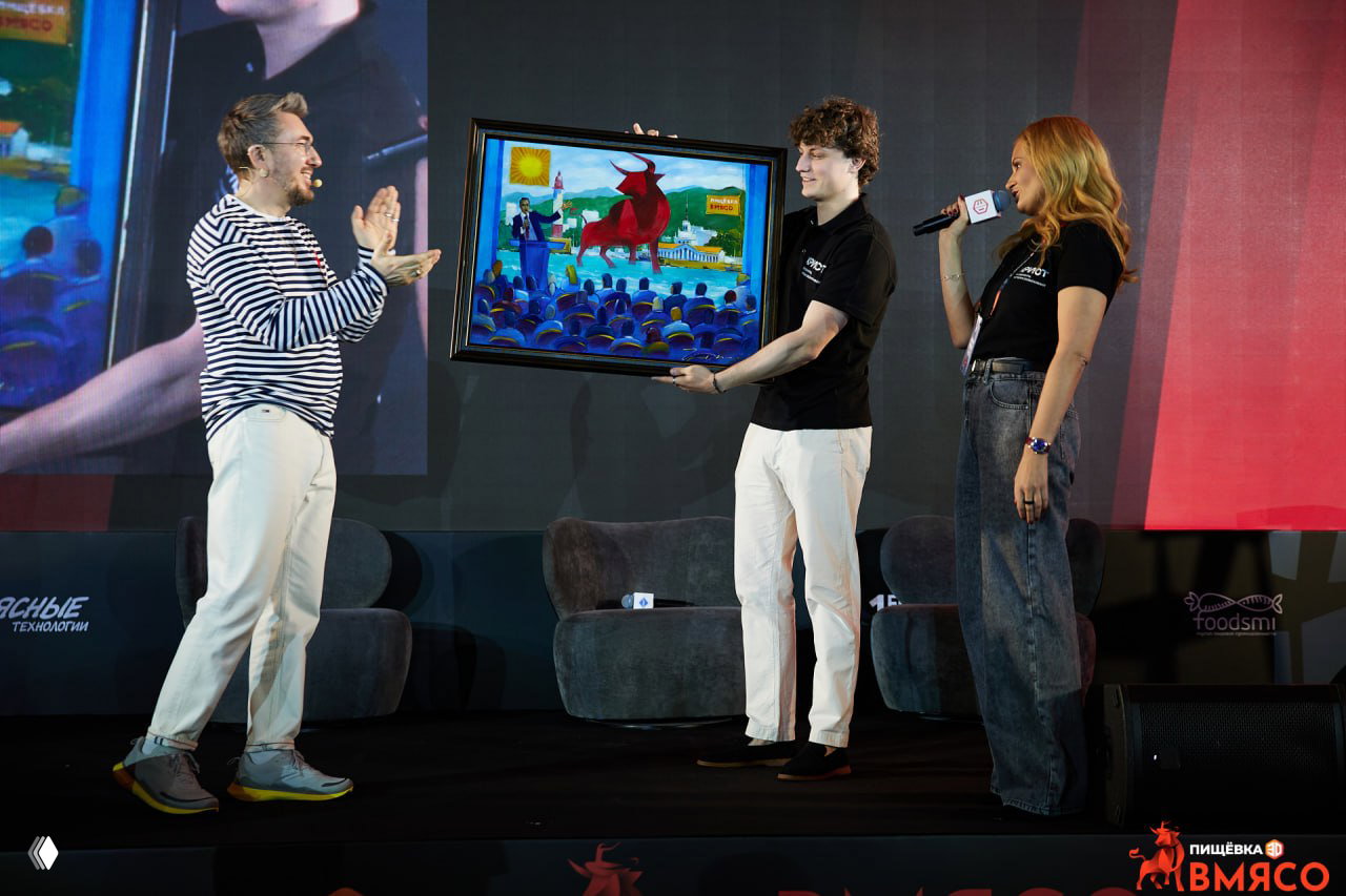 Церемония на сцене: вручение картины в рамках программы ВМЯСО, ведущие и участник держат художественное полотно перед аплодирующей аудиторией.