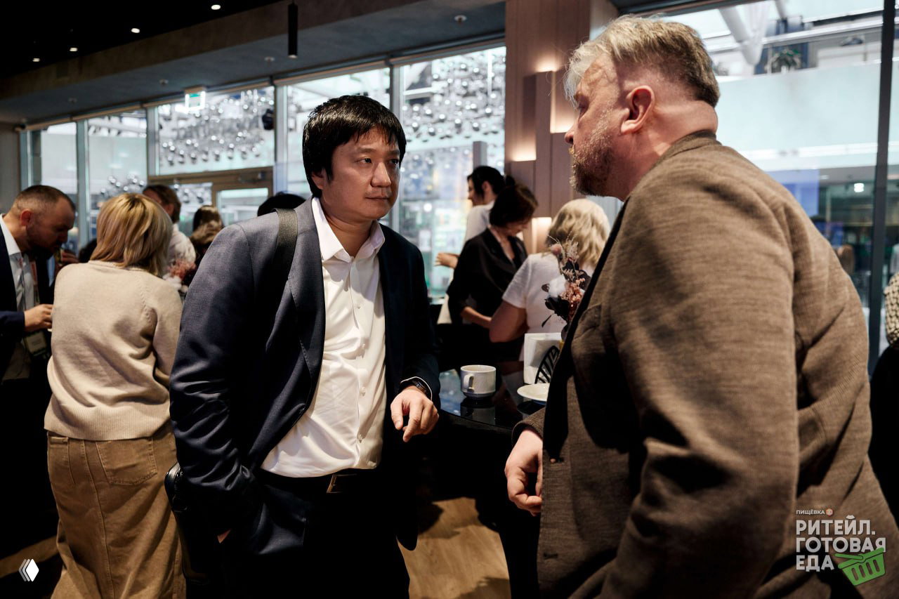 Двое участников ведут деловой разговор у стойки кофе-брейка на конференции по дистрибуции готовой еды; вокруг другие гости и зона нетворкинга.