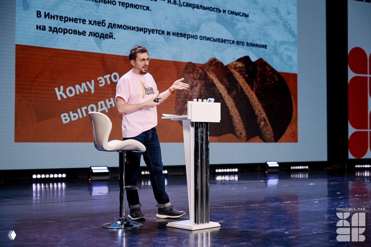 Докладчик на сцене Пищёвка.ЛАБ: спикер в розовой футболке выступает на фоне слайда с изображением хлеба и ключевых тезисов.