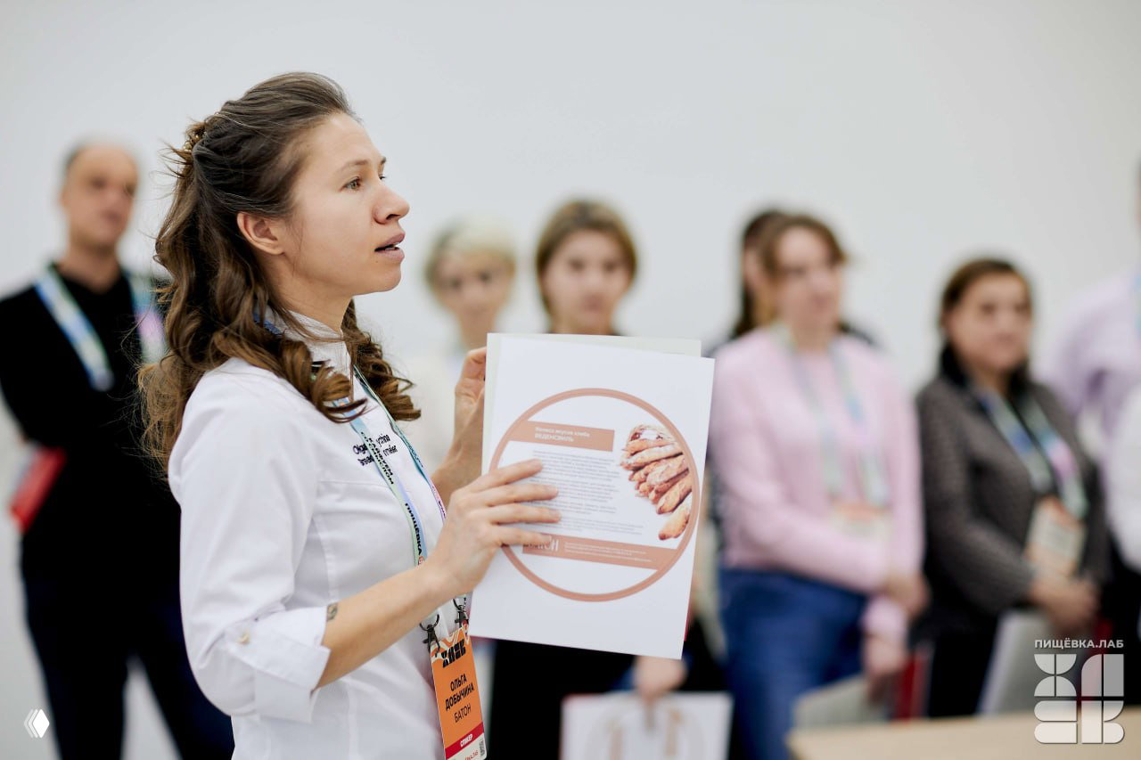 Женщина-лектор демонстрирует раздаточный материал о хлебе участникам лаборатории: крупный план, слушатели на заднем плане.