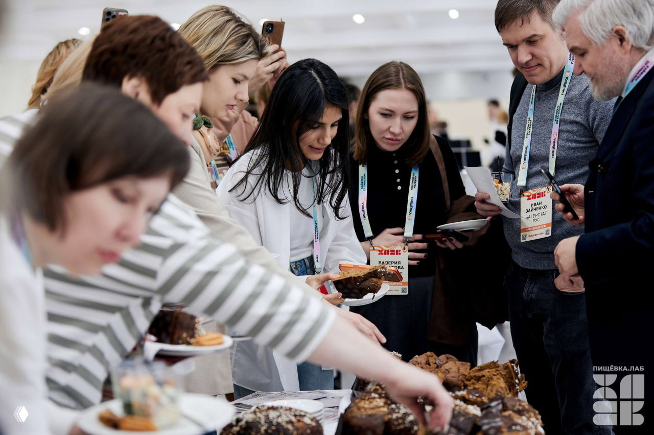 Посетители дегустируют хлебобулочные изделия на столе фуршета Пищёвка.ЛАБ: люди пробуют и обсуждают образцы, масштабная подача.