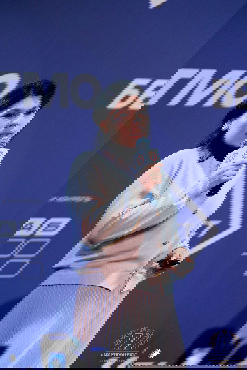 Крупный план: женщина-спикер держит микрофон на фоне брендированного баннера Ассортиматики, момент выступления.