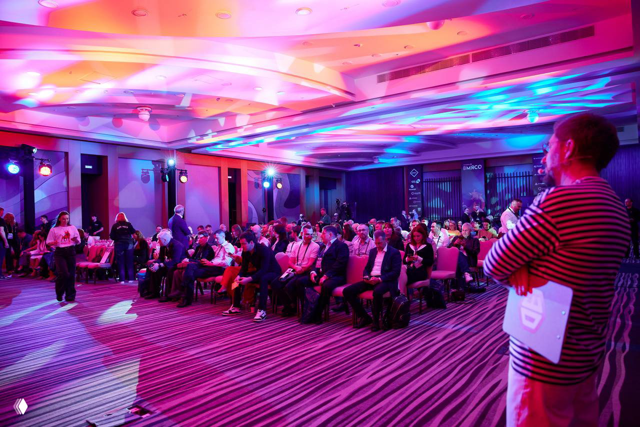 Широкий план зала с красочными сценическими огнями: участники сидят в рядах, фиолетово-розовая подсветка создает атмосферу события.