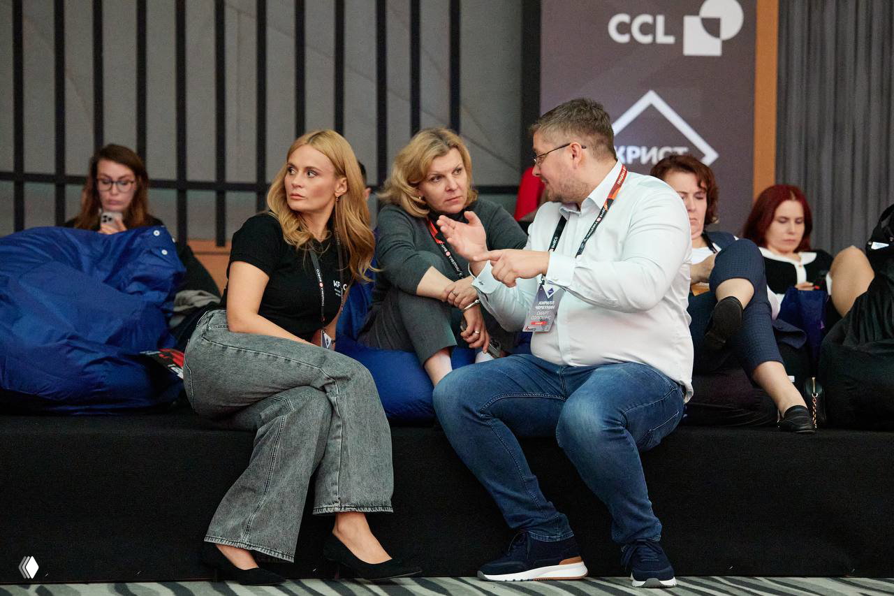 Небольшая группа участников ведет оживленную беседу, сидя на пуфах у сцены: живой диалог и обмен мнениями в перерыве мероприятия.