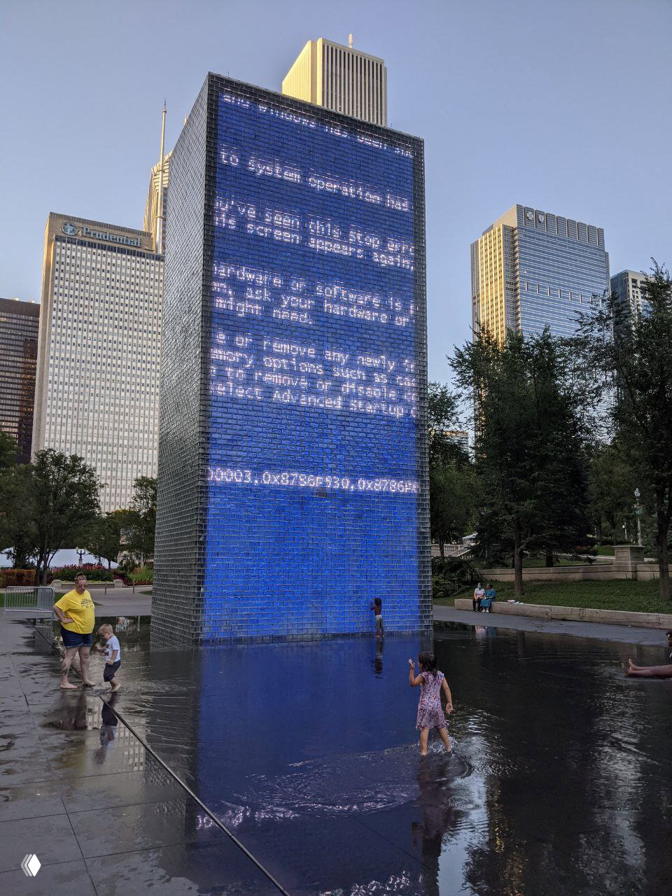Вертикальная панель Crown Fountain вблизи на фоне чикагских небоскрёбов: экран показывает ошибку BSOD, дети ходят по мокрой площади перед фонтаном.